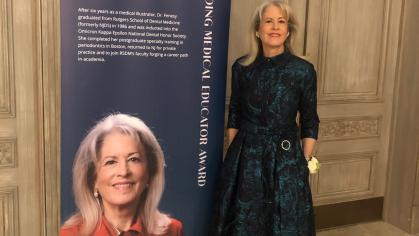  Dr. Kim E. Fenesy stands beside a tall banner displaying her photo and biography at the 2025 Excellence in Medicine Awards. She is wearing a long, dark teal and black formal gown with a floral pattern. The banner recognizes her as the recipient of the Outstanding Medical Educator Award and includes a professional portrait of her in a red blazer. The ornate setting suggests a formal awards ceremony.