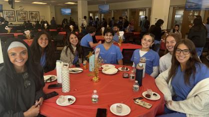 "A group of people, wearing blue scrubs, are seated around a table with a red tablecloth. The table holds plates of food, water bottles, and phones. In the background, more tables and people are visible.