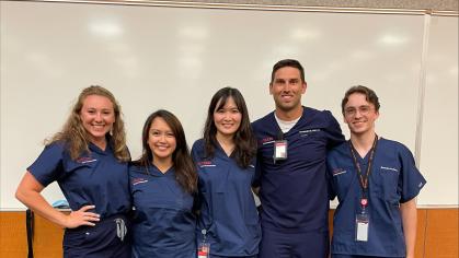 A group of five students wearing navy blue scrubs stand together in a classroom setting. They are positioned in front of a whiteboard. 