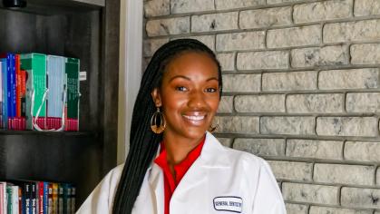 A person with long braided hair stands in front of a bookshelf and a brick wall, wearing a white coat over red scrubs. 