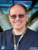 A man wearing glasses and dark scrubs with a name badge smiles in front of a modern glass building.