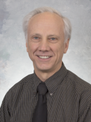 Smiling older man with short white hair wearing a black-and-white checkered shirt and a black tie, in front of a light gray mottled background.
