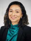 Professional headshot of a woman with wavy, shoulder-length dark hair and subtle highlights, wearing a teal satin blouse, black blazer, and pearl earrings, smiling against a plain light gray background.