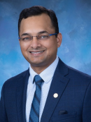 Professional headshot of a smiling man wearing glasses, a dark blue suit, a blue patterned tie, and a light blue checkered shirt, standing against a mottled blue background.