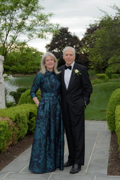 Kim Fenesy and George Heinrich stand side by side on a stone pathway in a manicured garden with lush greenery and a pond in the background. Fenesy wears a long, elegant dark teal and black gown with a floral pattern, while Heinrich is dressed in a black tuxedo with a bow tie and white rose boutonnière. They are smiling and appear dressed for the award ceremony.