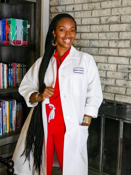 A person with long braided hair stands in front of a bookshelf and a brick wall, wearing a white coat over red scrubs. 